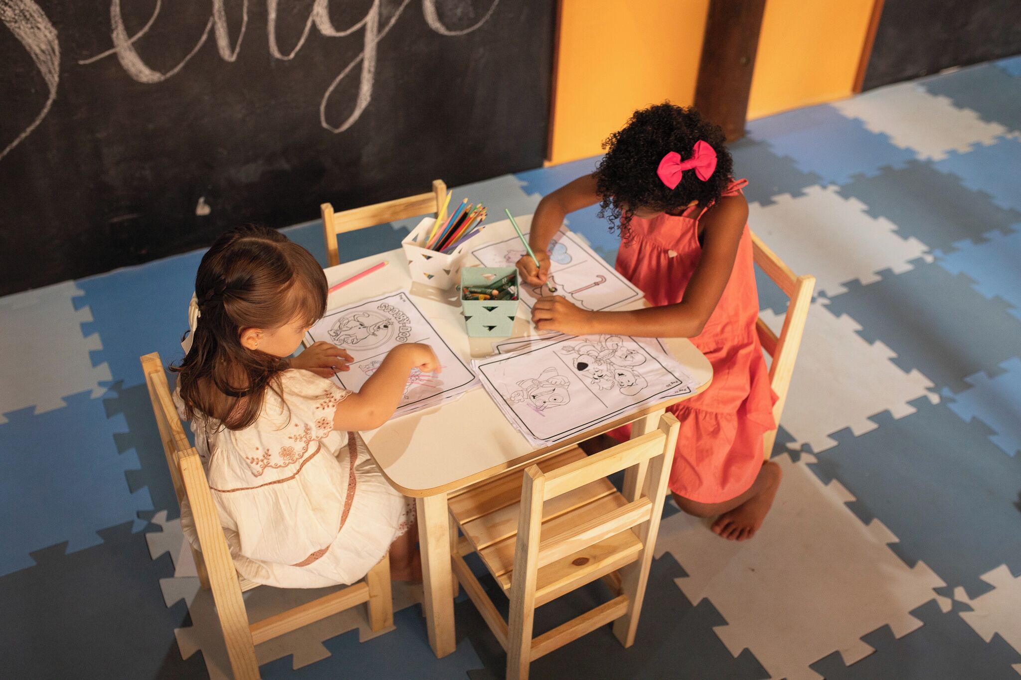 Dos niños dibujando en una mesa con una pizarra de fondo.