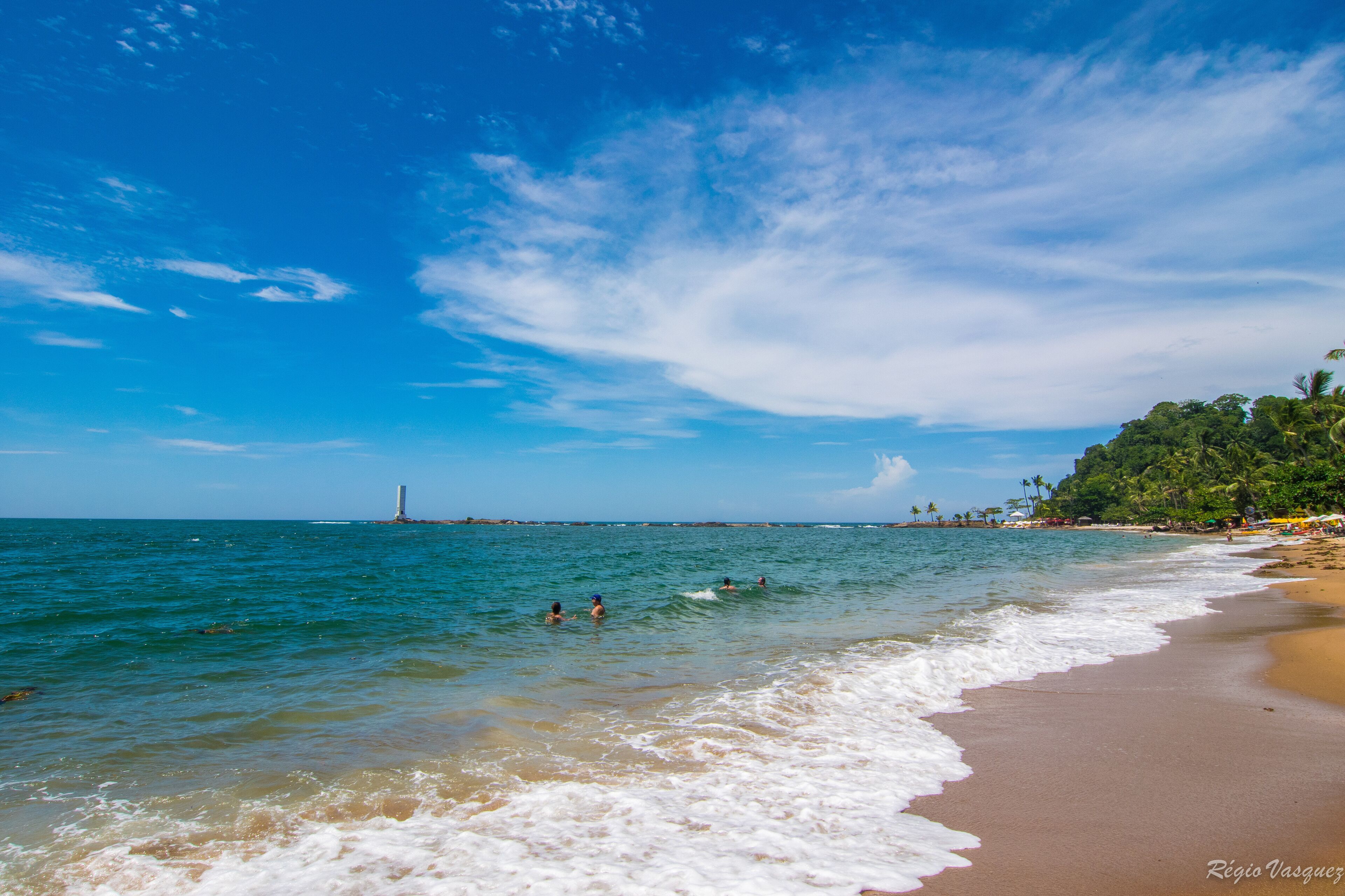 A beach with clear blue water, a sandy shore, and a lighthouse in the distance.