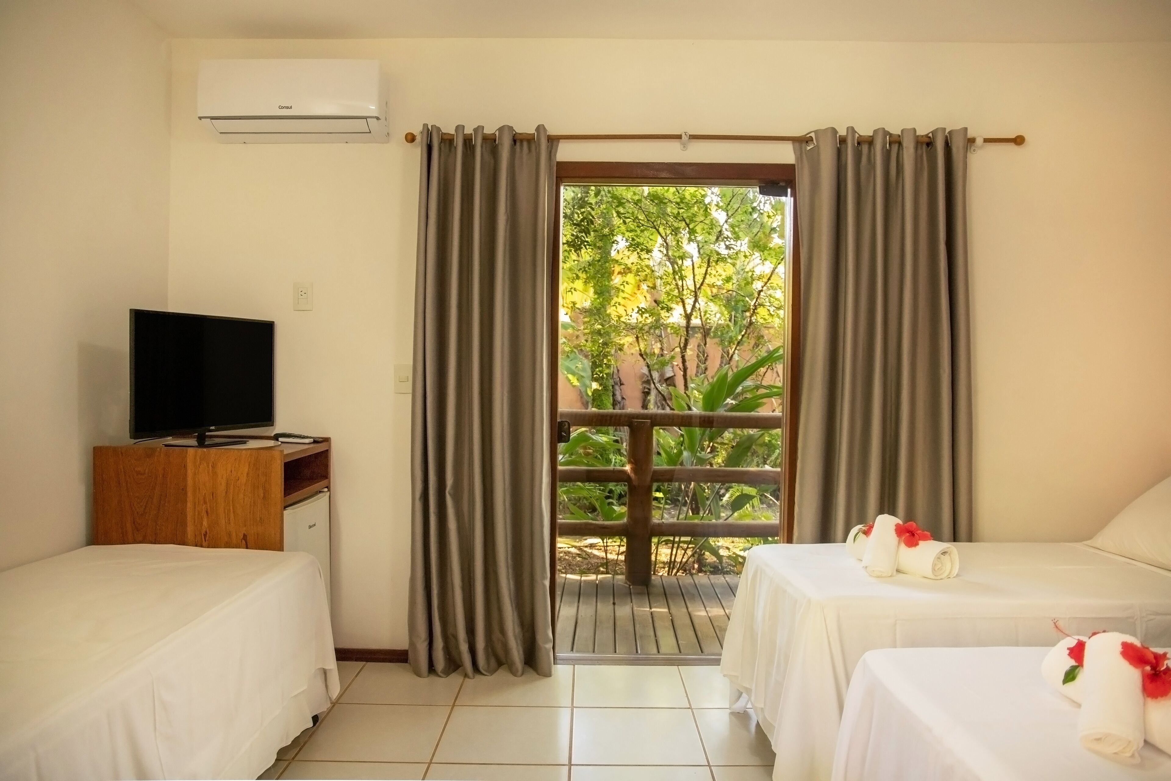A hotel room with two beds, a TV on a wooden cabinet, and a view of greenery through a large window.