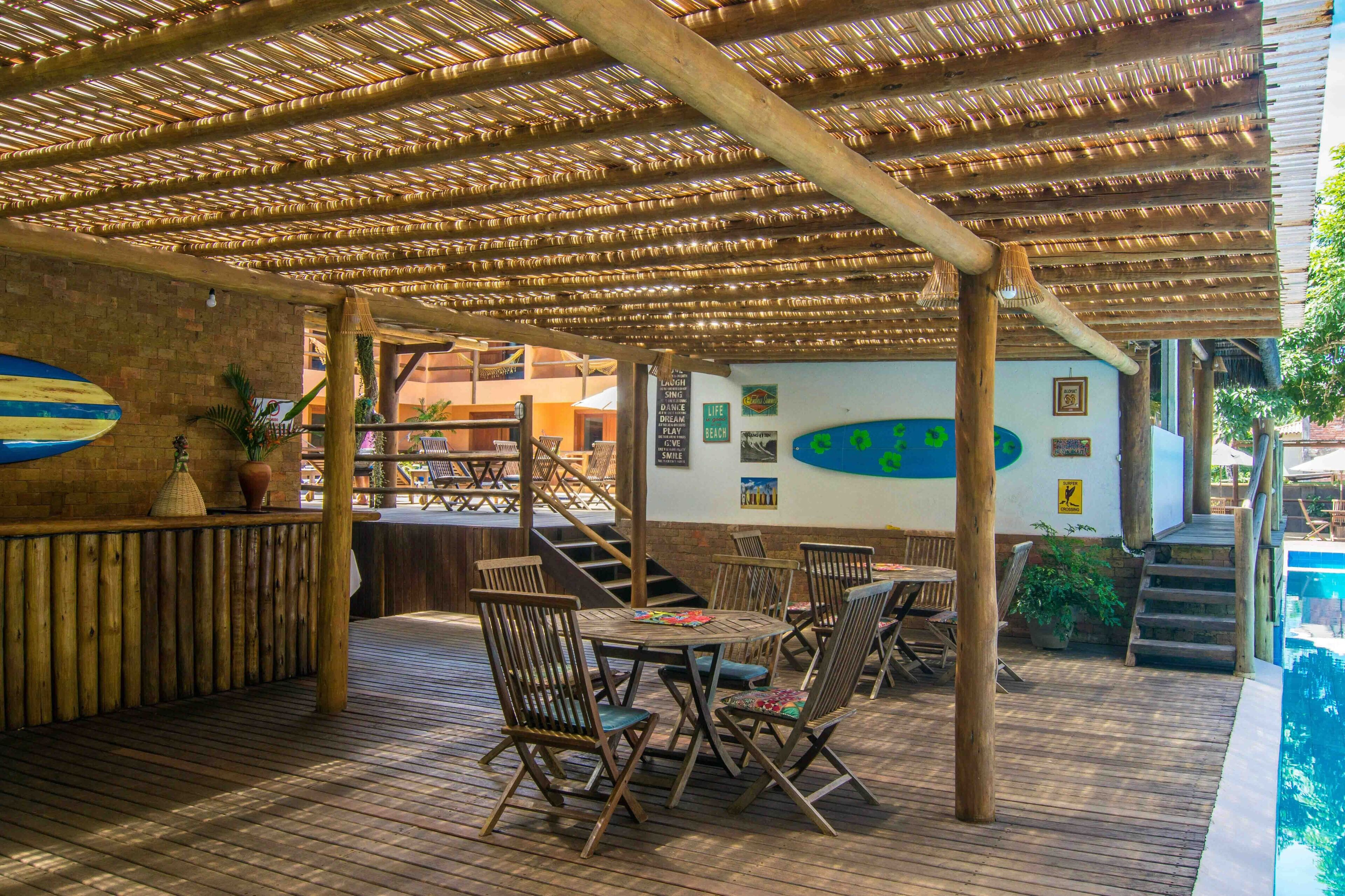 Un espacio al aire libre con mesas y sillas de madera, una piscina y una zona de estar techada con techo de bambú.