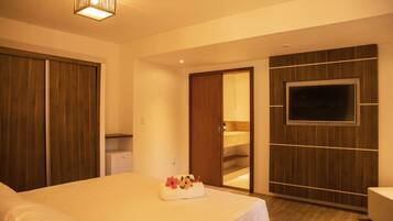 Habitación de hotel con cama, televisor montado en la pared, armario de madera y pequeña cocina.