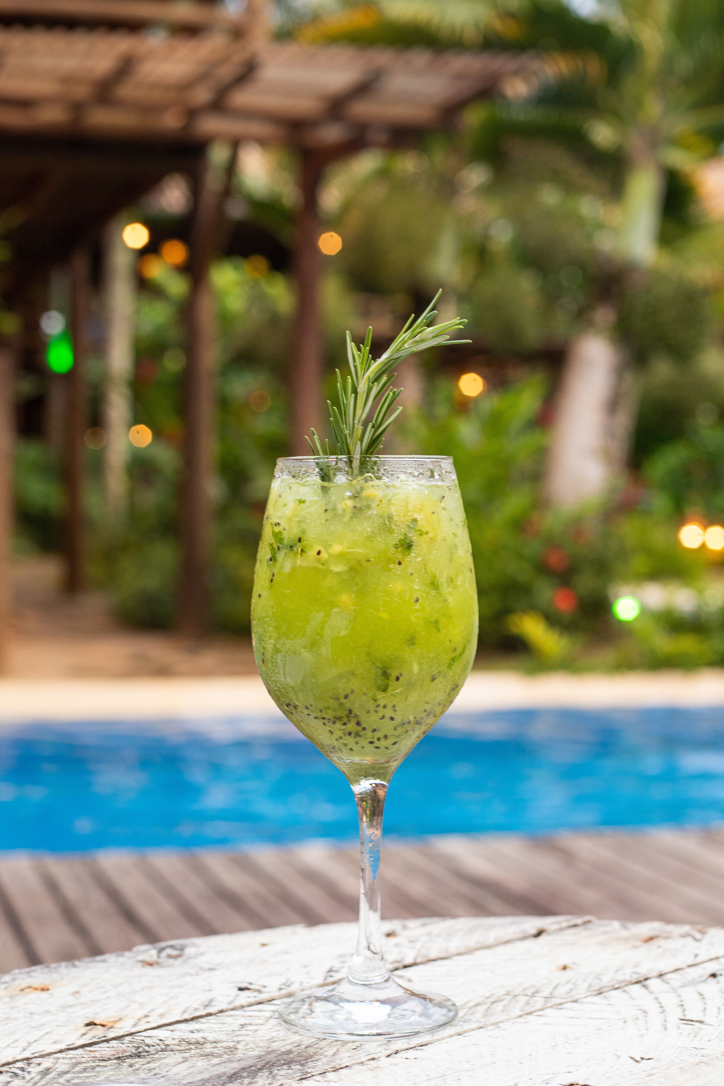 A cocktail glass with a sprig of rosemary on a wooden table by a pool.