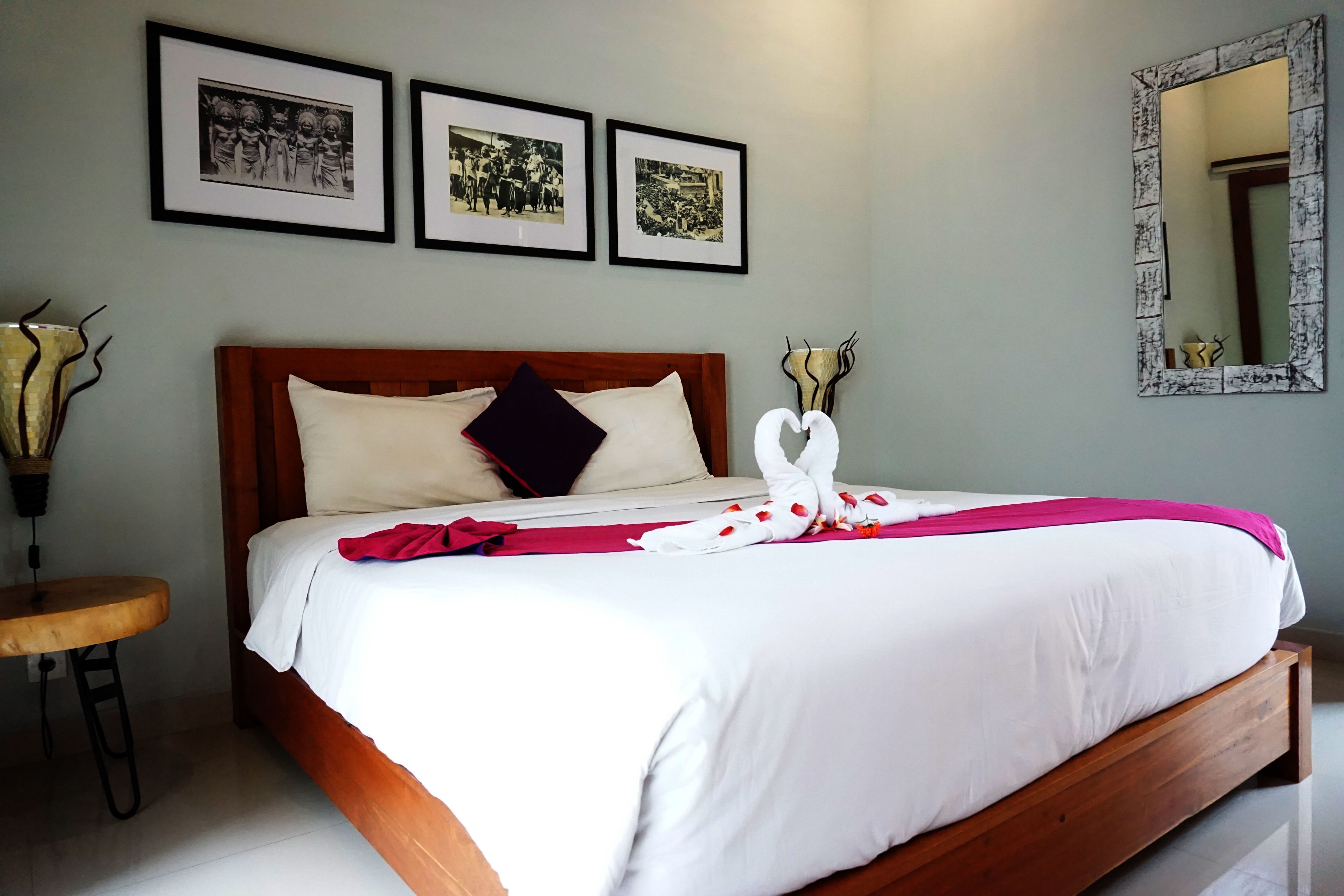 A neatly made bed with a wooden headboard, white bedding, and a red and white decorative pillow. There are three framed pictures on the wall above the bed.