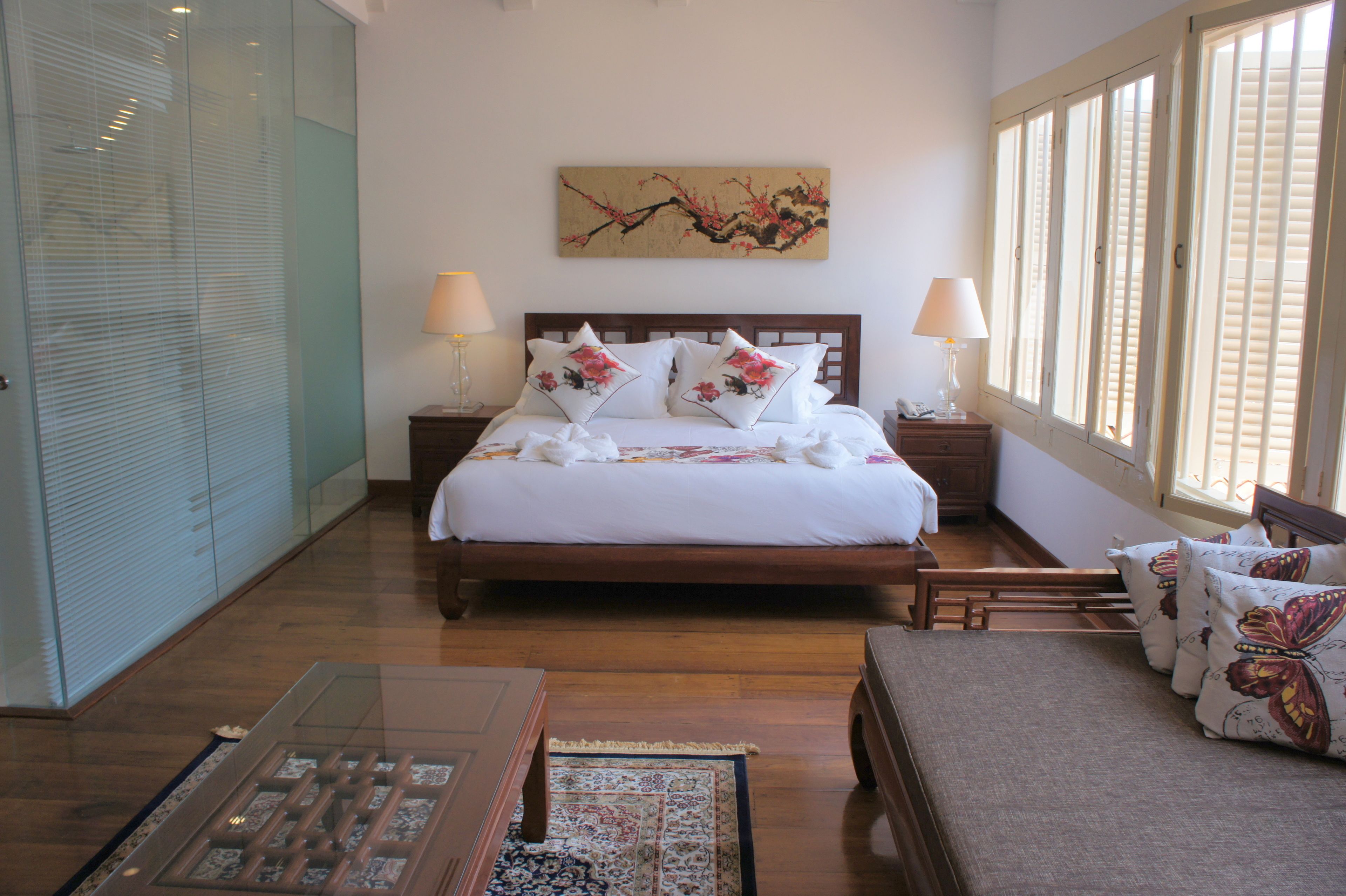 A neatly arranged bedroom with a wooden bed, two bedside tables with lamps, a large window with blinds, and a decorative wall piece.
