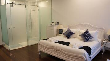 A hotel room with a glass-enclosed shower, a white bed with two pillows, a bedside table with a lamp, and a small nightstand with a phone.