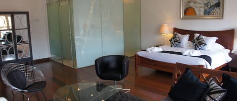 A hotel room with a bed, a glass table, a black chair, a sofa, a wooden bench, and a glass-enclosed wardrobe.