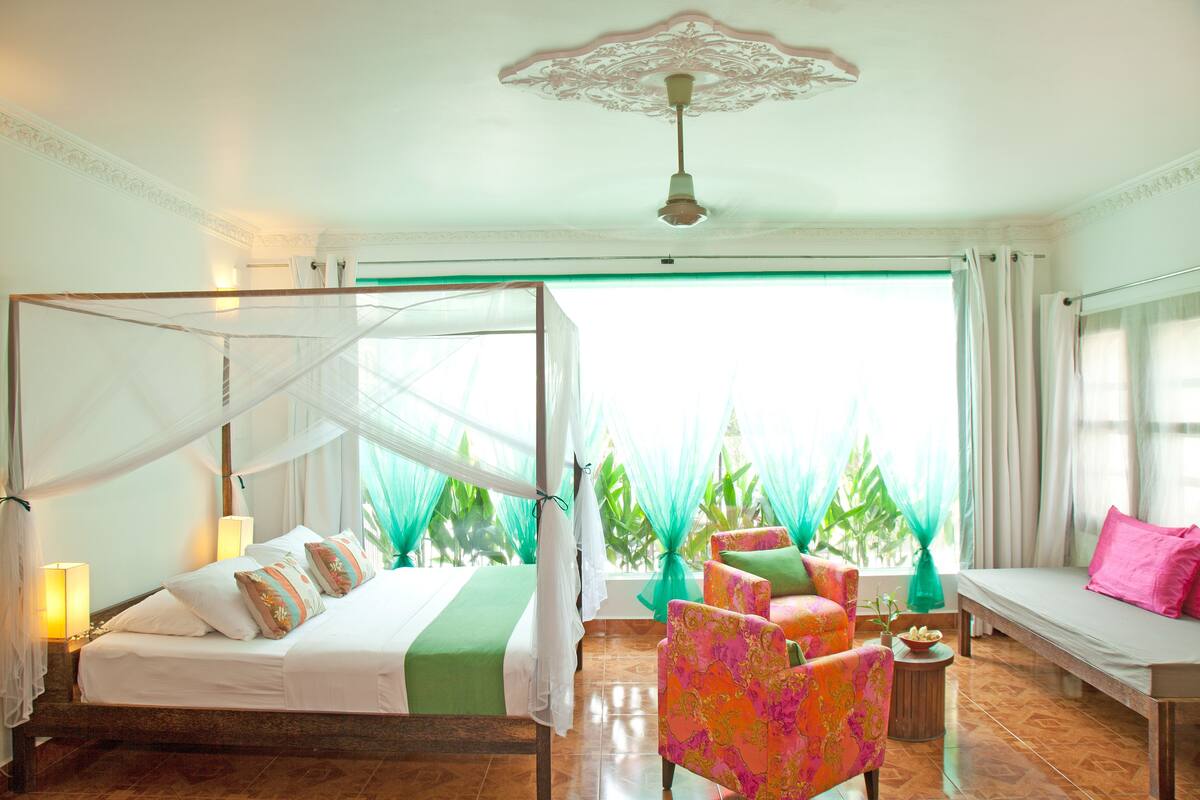 A room with a bed, a bench, and a chair. There is a window with curtains and a view of greenery outside.