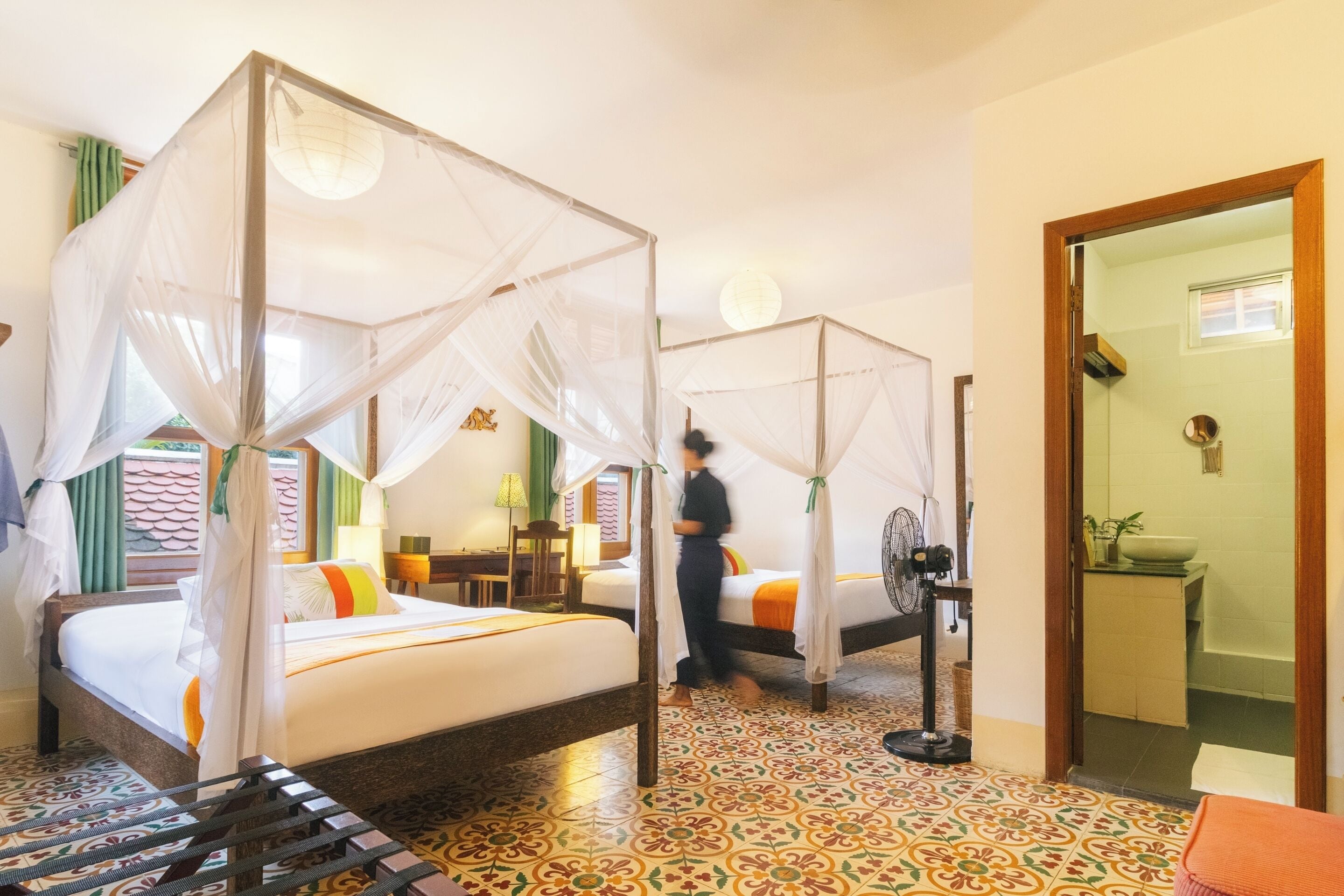 A hotel room with two beds, a standing fan, a bathroom visible through an open door, and a patterned floor.
