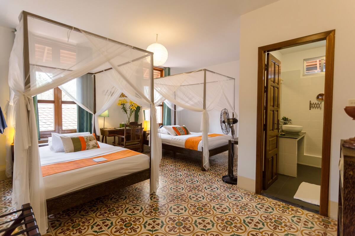 A room with two beds, each with a canopy, a tiled floor, a bedside table, and a window with curtains.