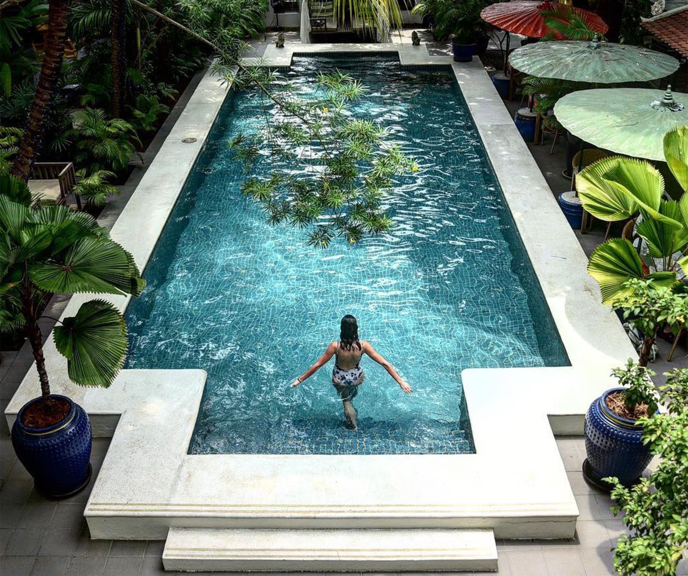 A rectangular pool with clear water, surrounded by potted plants and umbrellas.
