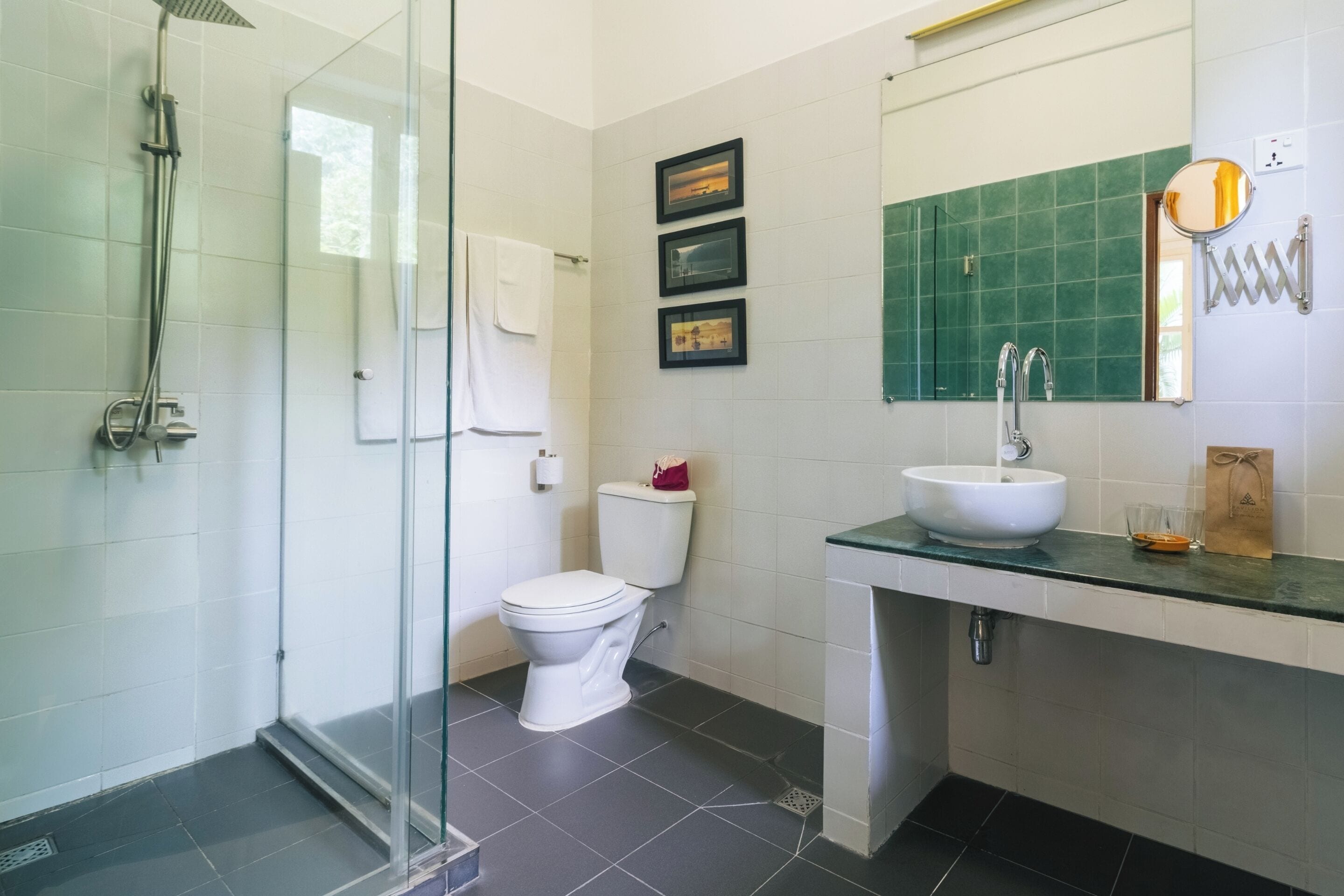 A modern bathroom with a glass shower enclosure, a toilet, a sink with a green countertop, and framed pictures on the wall.