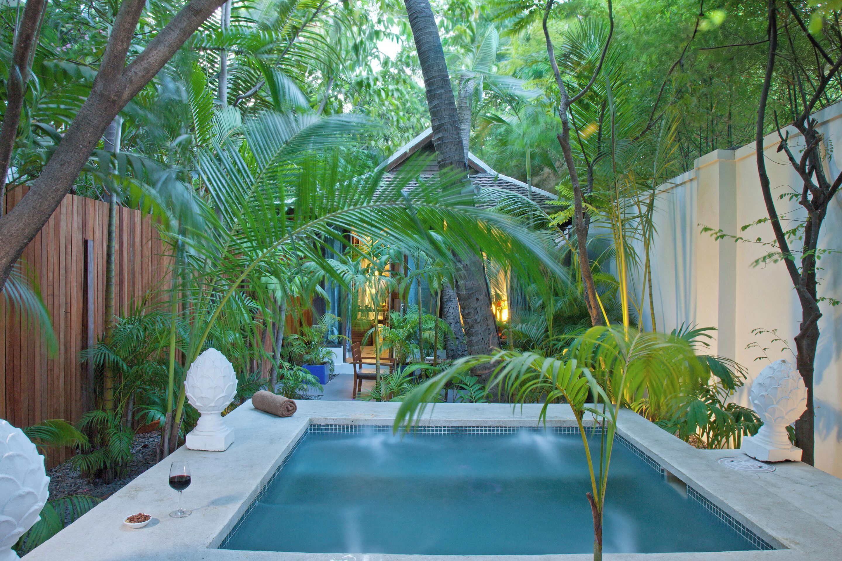 Una piscina rodeada de vegetación exuberante, una valla de madera y esculturas blancas decorativas.
