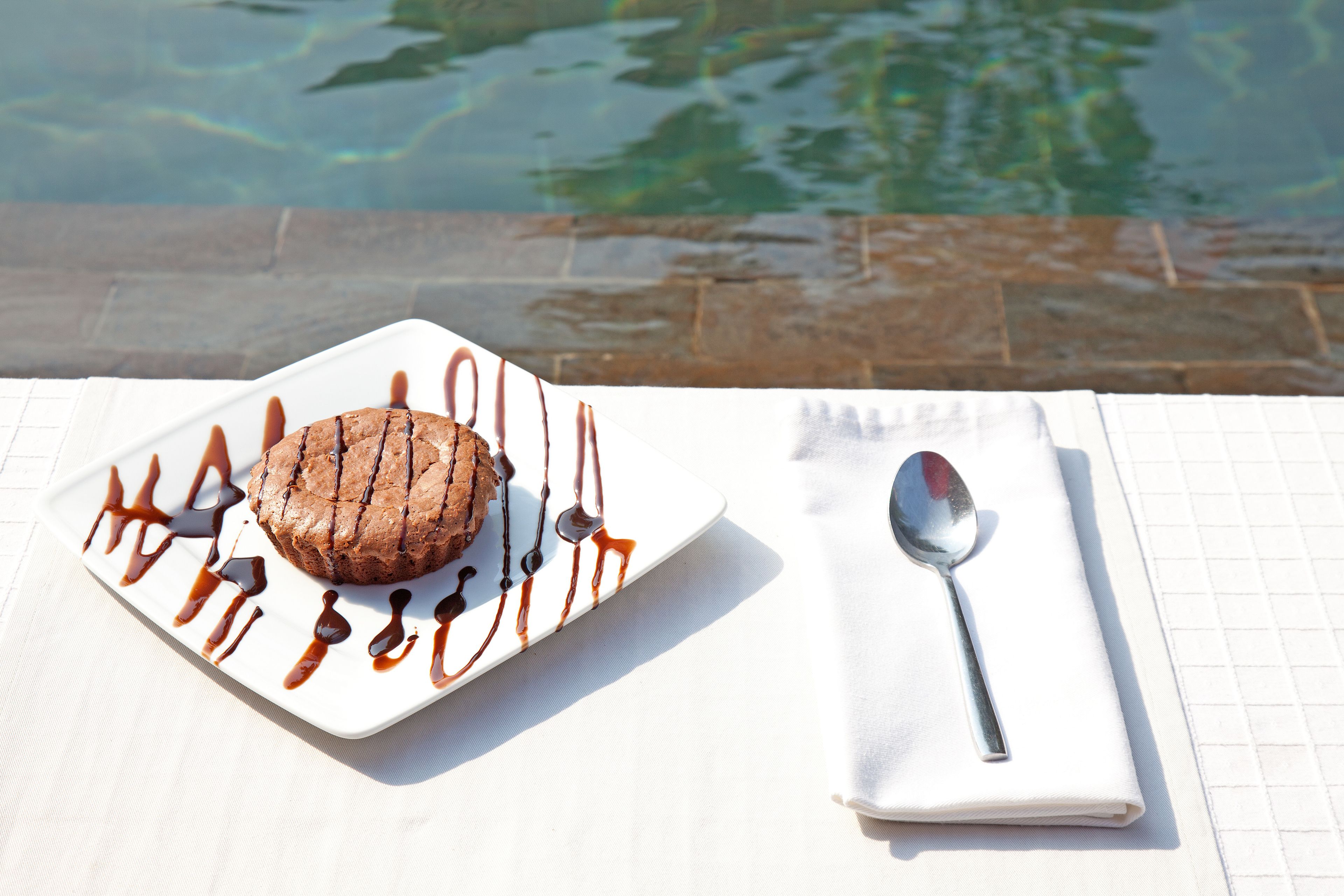Postre sobre un plato blanco con un chorrito de chocolate, acompañado de una cuchara y servilleta, colocado sobre una mesa cerca de una piscina.