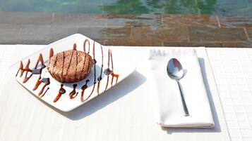 Postre sobre un plato blanco con un chorrito de chocolate, acompañado de una cuchara y servilleta, colocado sobre una mesa cerca de una piscina.