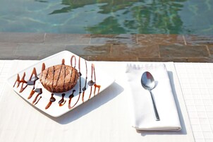 Postre sobre un plato blanco con un chorrito de chocolate, acompañado de una cuchara y servilleta, colocado sobre una mesa cerca de una piscina.