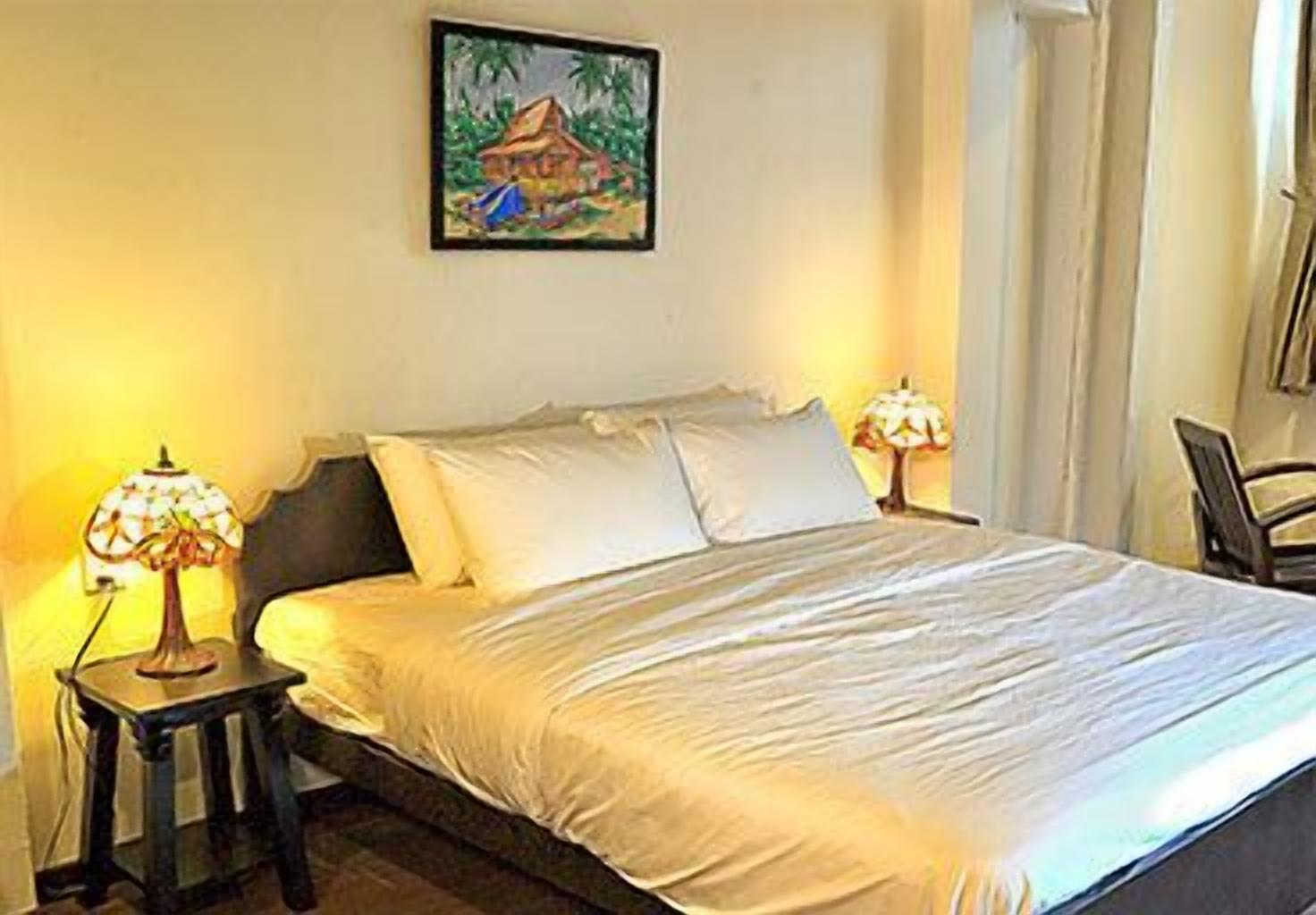 A neatly made bed with white linens, two bedside lamps, and a painting of a tropical scene on the wall.