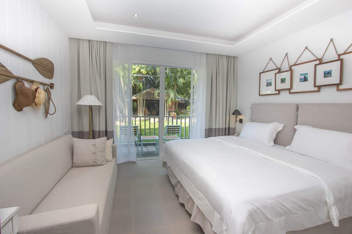 A modern hotel room with a large bed, a sofa, a balcony with chairs, and a view of greenery.