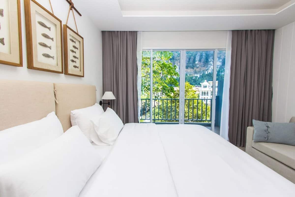 A hotel room with a large bed, a sofa, a balcony with a view of greenery and buildings, and framed pictures on the wall.