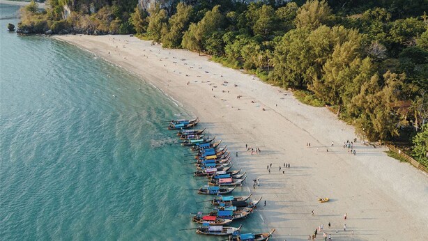 Een overzicht vanuit de lucht van een strand met aan de kust afgemeerd bootjes, een zandstrand en weelderige begroeiing op de achtergrond.