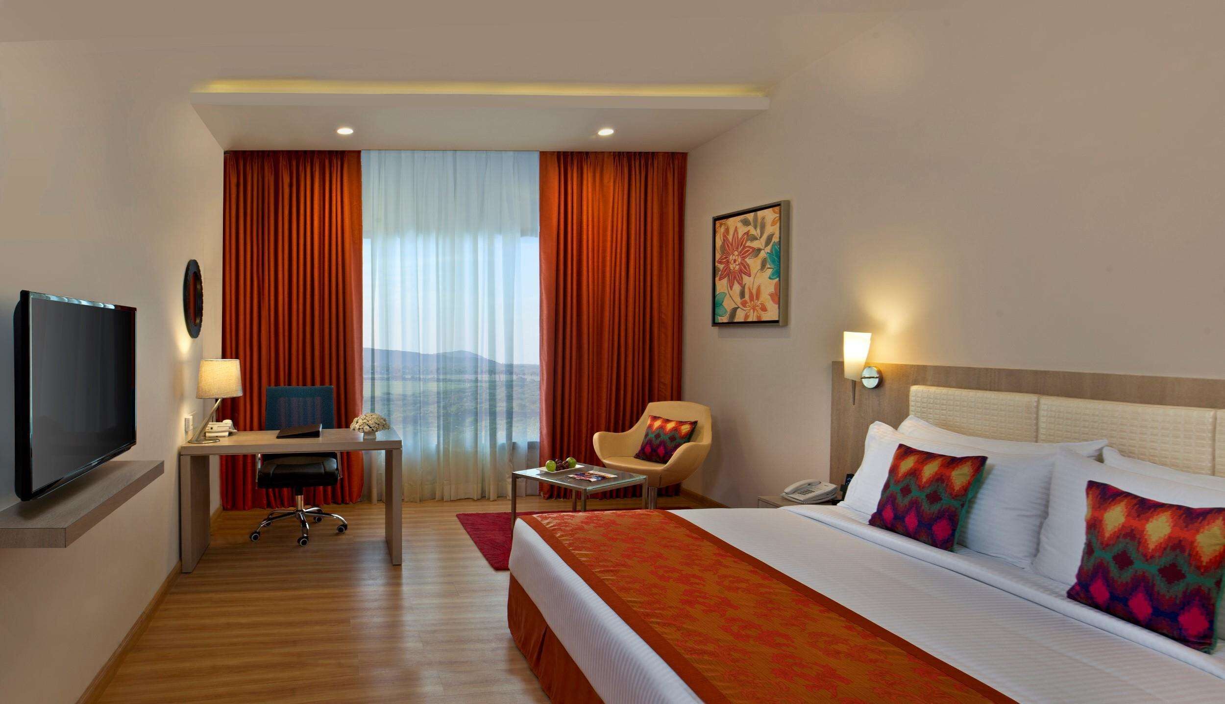 Habitación de hotel con cama, escritorio con computadora, silla, televisor y ventana con cortinas.
