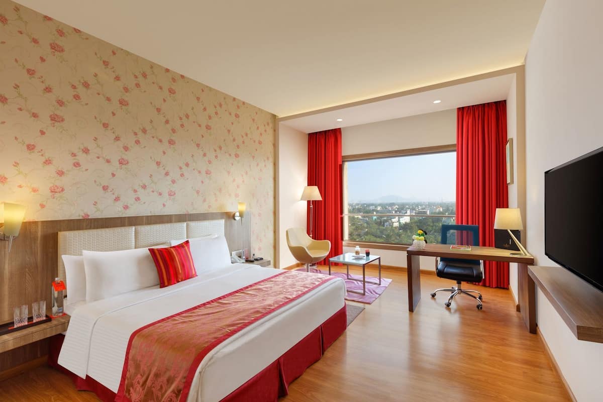 Habitación de hotel con cama, escritorio, silla, televisor y ventana grande con vista a la ciudad.
