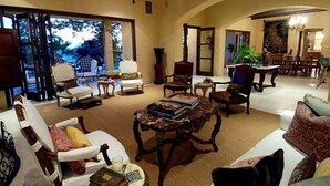 Amplia sala de estar con techo alto, ventanas grandes y vista a un jardín. Hay varias zonas de estar con sofás y sillones, además de un comedor con mesa y sillas.