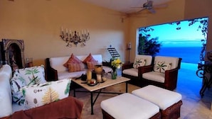 Un salón con sofá, sillones y otomanas, una mesa de centro con objetos decorativos, un candelabro y vista al océano.