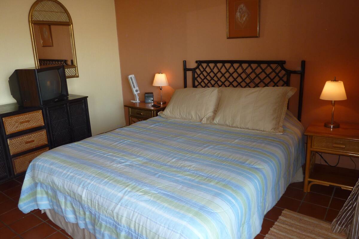 A bedroom with a large bed, two bedside lamps, a nightstand, a dresser, a television, and a mirror.