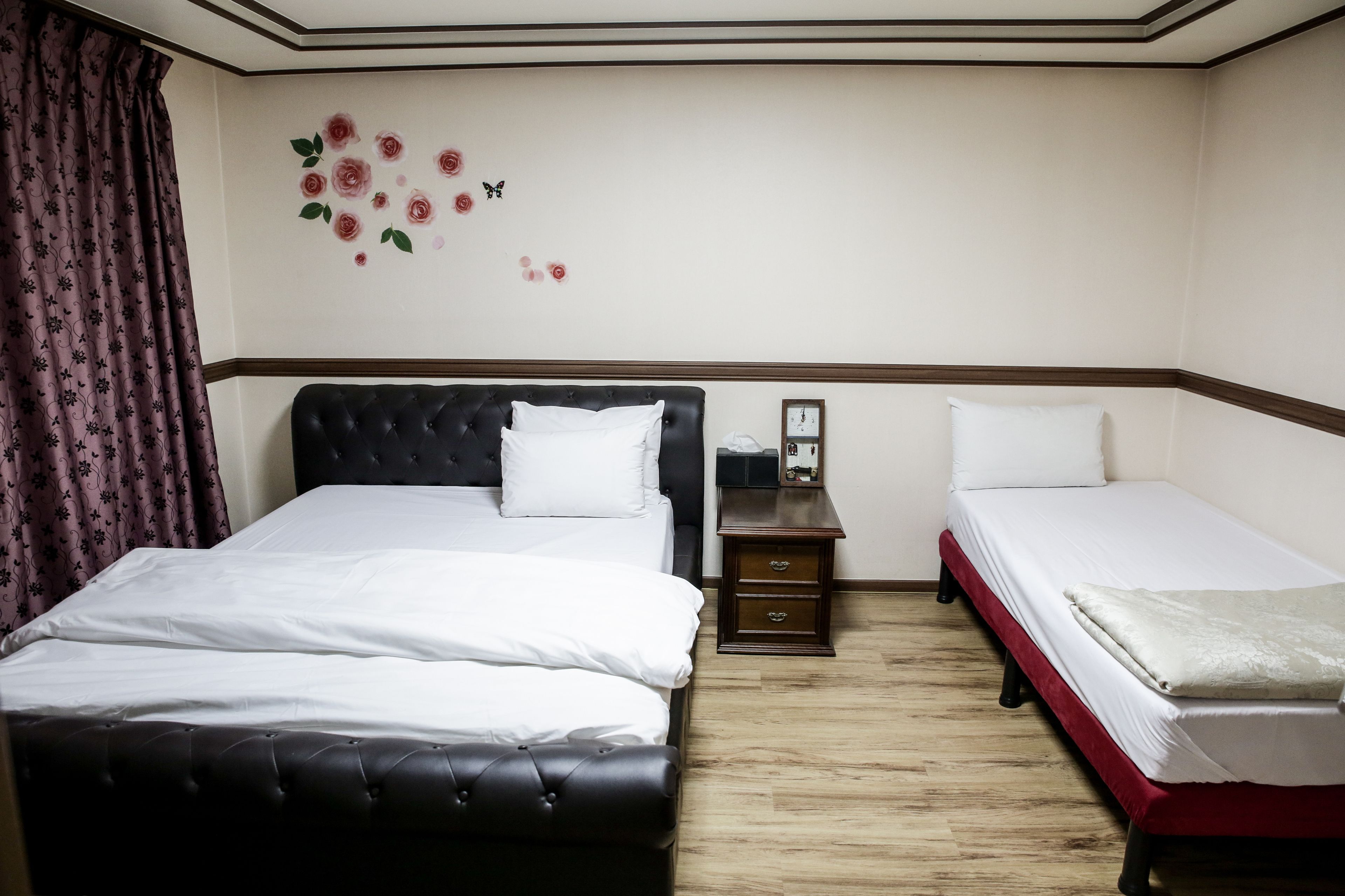 Habitación de hotel con dos camas, una con cabecera negra y la otra con cabecera roja. Entre las camas hay una mesita de noche de madera con un reloj y un teléfono. Las paredes tienen adhesivos florales y el piso es de madera.