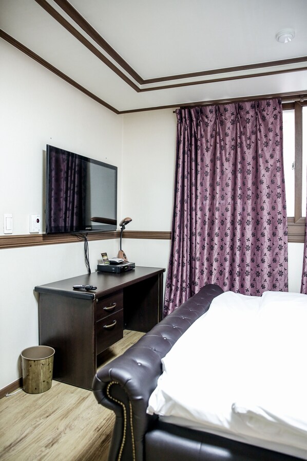 A hotel room with a bed, a desk with a flat-screen TV, a lamp, and a window with purple curtains.