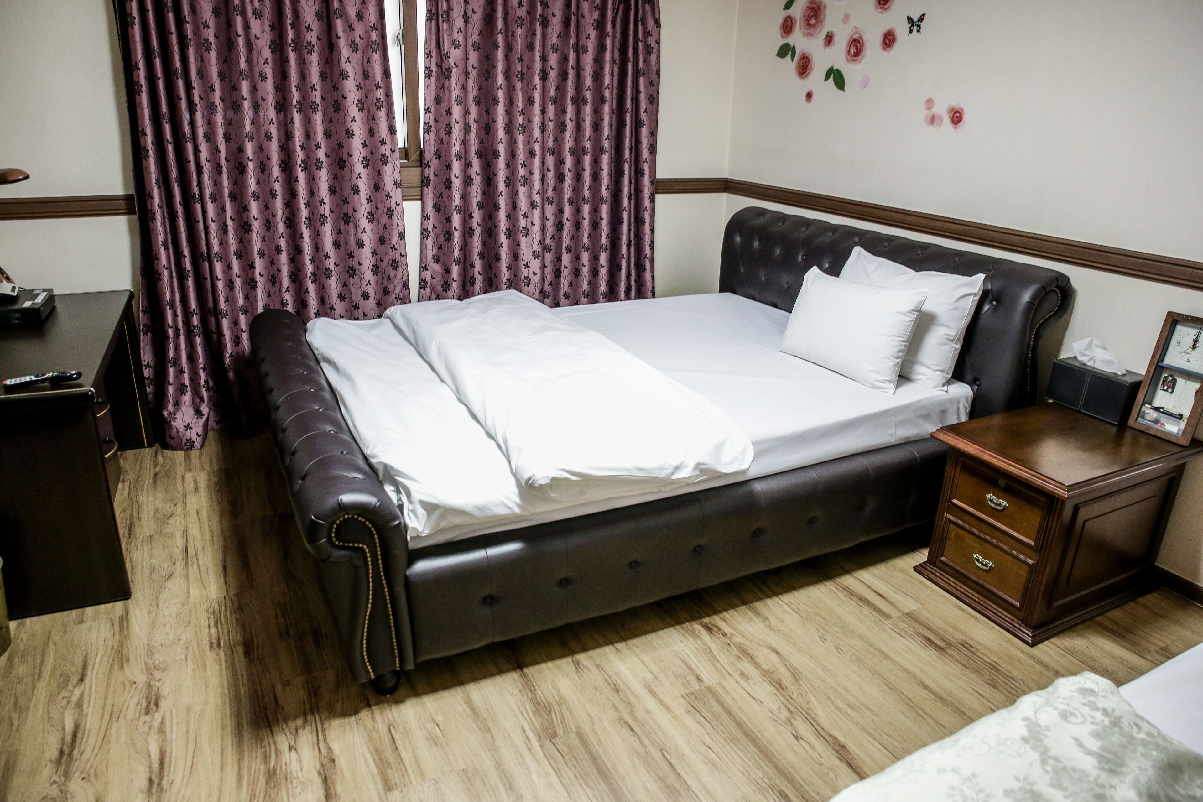 Una cama bien hecha con un cabecero oscuro, una mesita de noche de madera con cajones y una ventana con cortinas moradas.