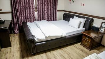 Una cama bien hecha con un cabecero oscuro, una mesita de noche de madera con cajones y una ventana con cortinas moradas.