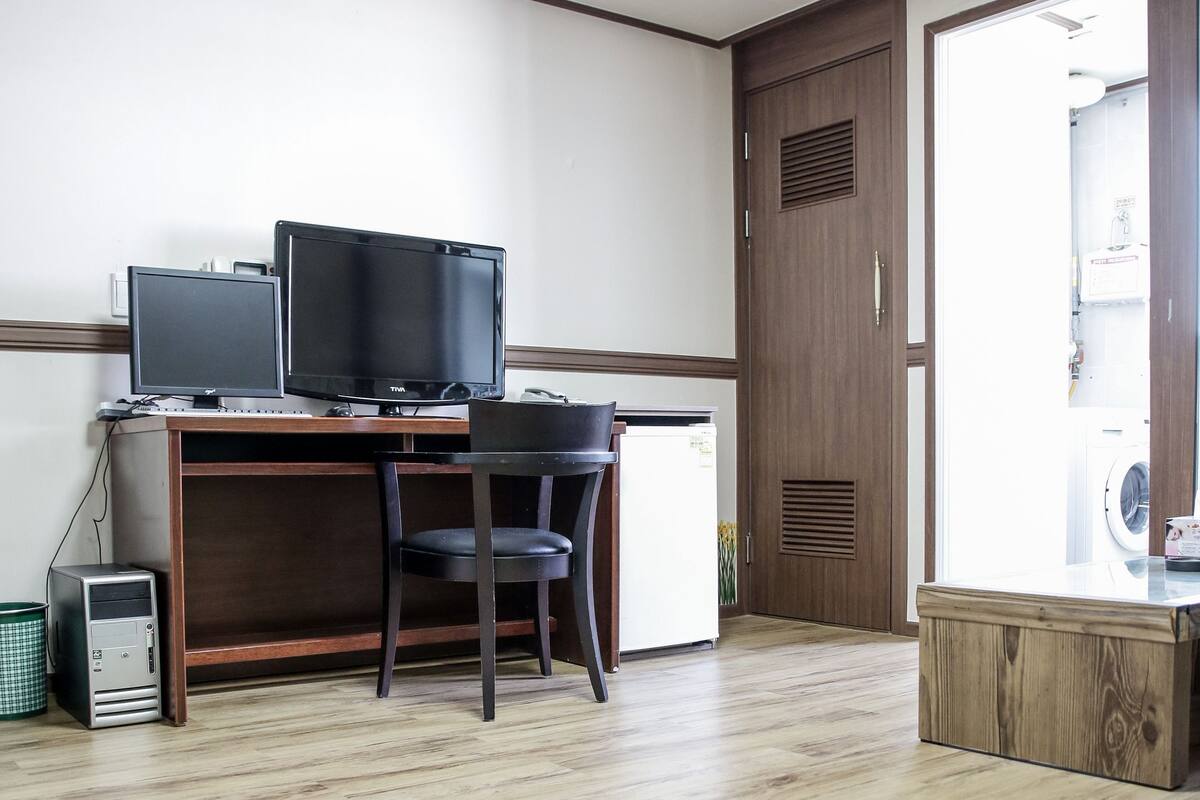 A room with a wooden desk, two monitors, a chair, a small refrigerator, and a washing machine.