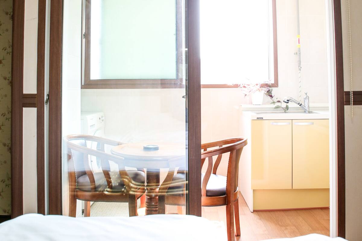 A room with a bed, a wooden chair, a small table, a window, and a kitchen area with cabinets and a sink.