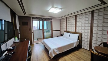Habitación de hotel con cama, escritorio con computadora, televisor y baño visible a través de una puerta de cristal.