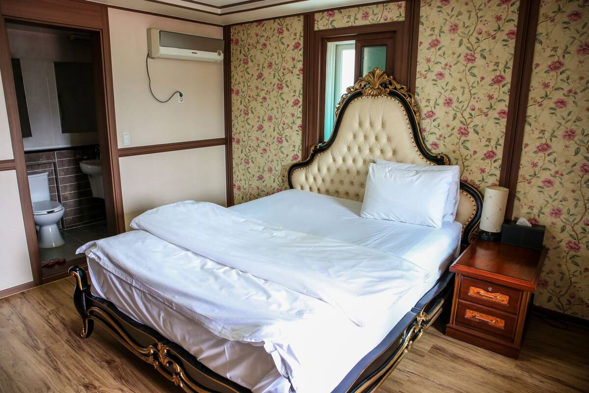 A bedroom with a bed, a nightstand, a mirror, and a bathroom visible through an open door.