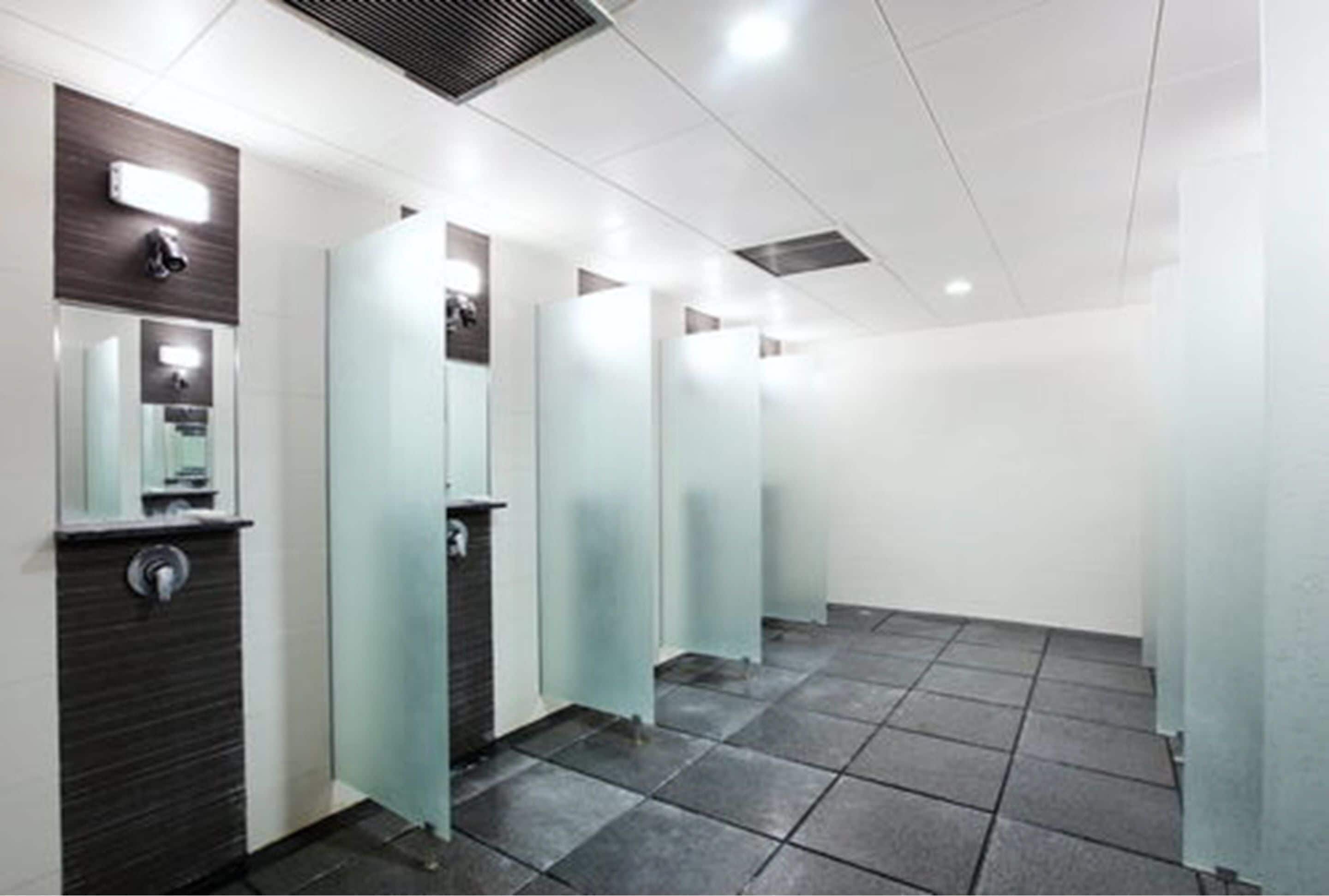 A row of modern, well-lit public restrooms with glass partitions and a tiled floor.