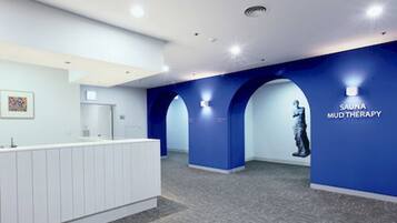 A modern spa reception area with a white reception desk, blue walls, and a sign for 'Sauna Mud Therapy'.