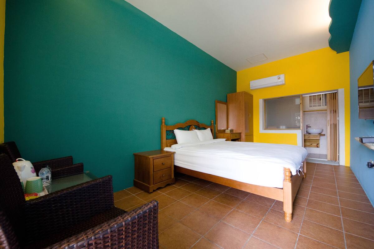 Un dormitorio con cama, mesitas de noche, una silla de mimbre y una pequeña mesa con una taza y una botella.