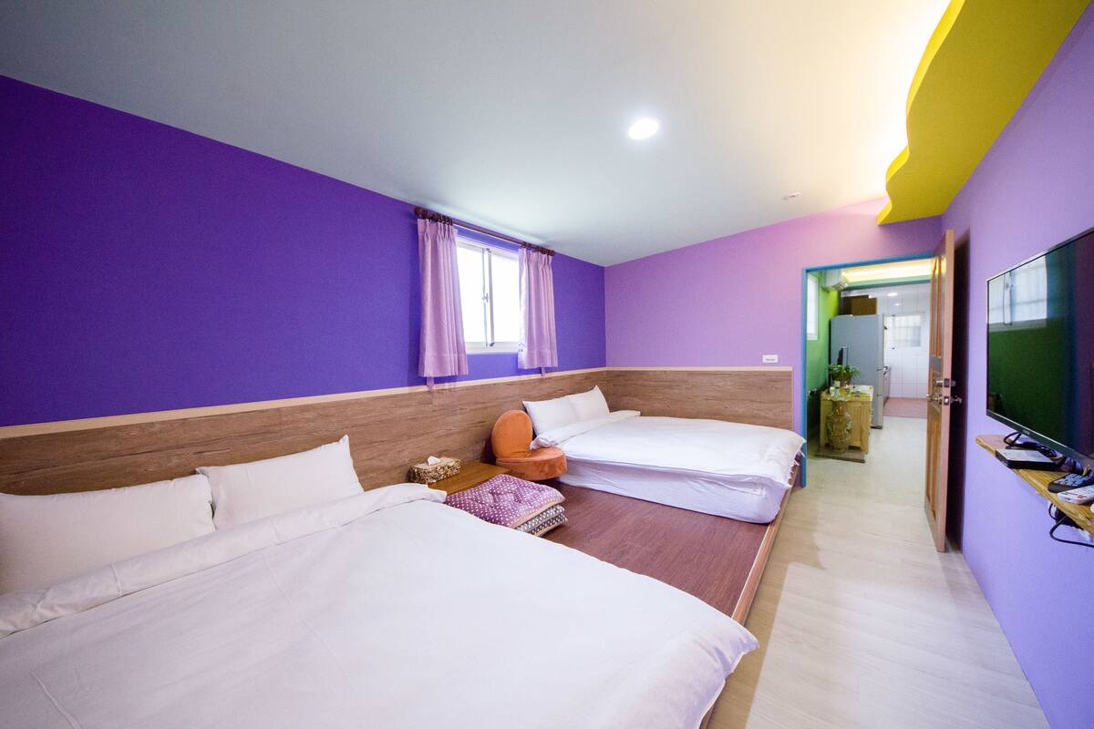 Habitación de hotel con dos camas, cabecera de madera, una silla naranja pequeña, televisor y baño visible a través de una puerta abierta.