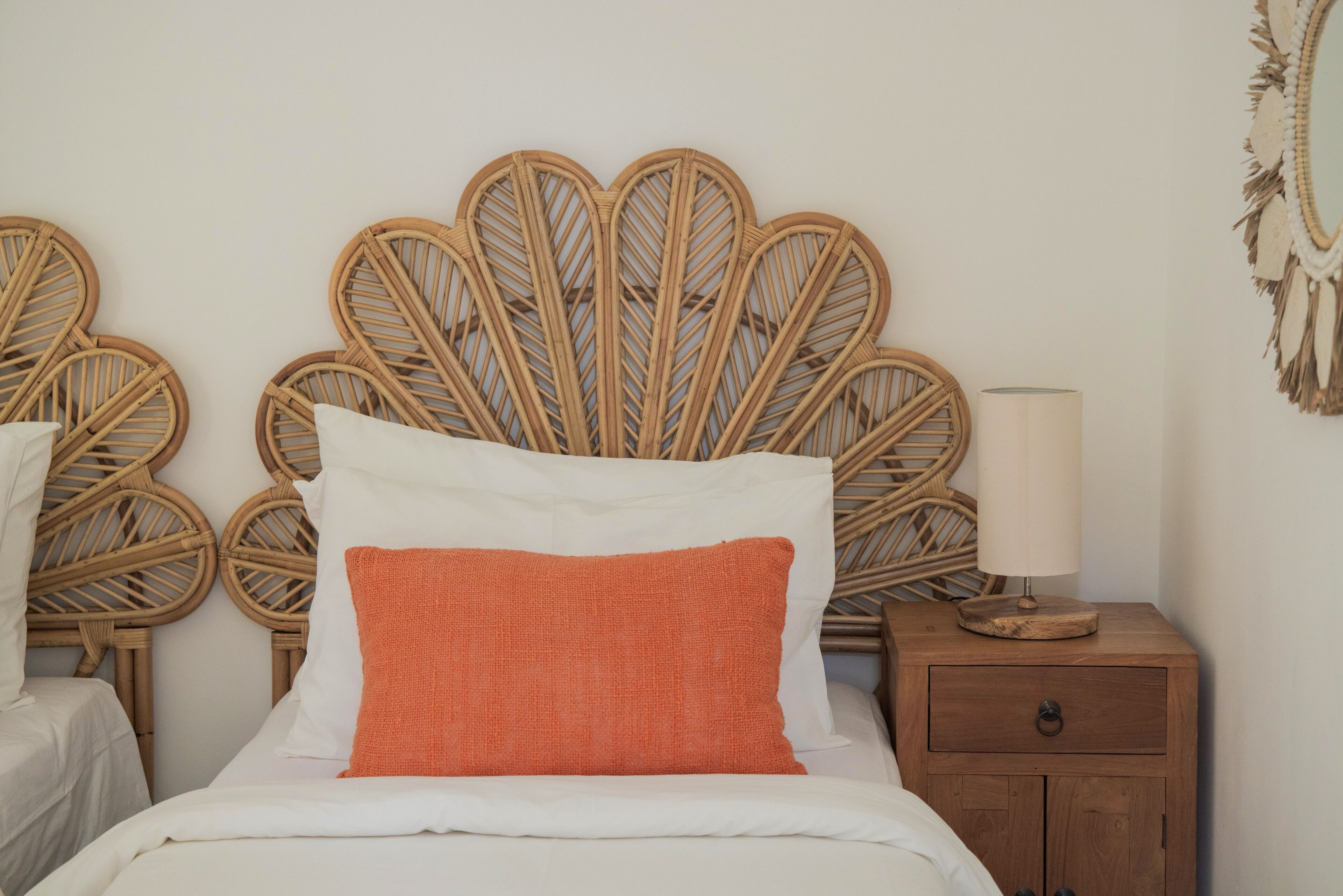 Une chambre avec un lit doté d’une tête de lit en rotin, de draps blancs et d’un oreiller orange. On y trouve une table de chevet en bois avec une lampe et un tiroir.