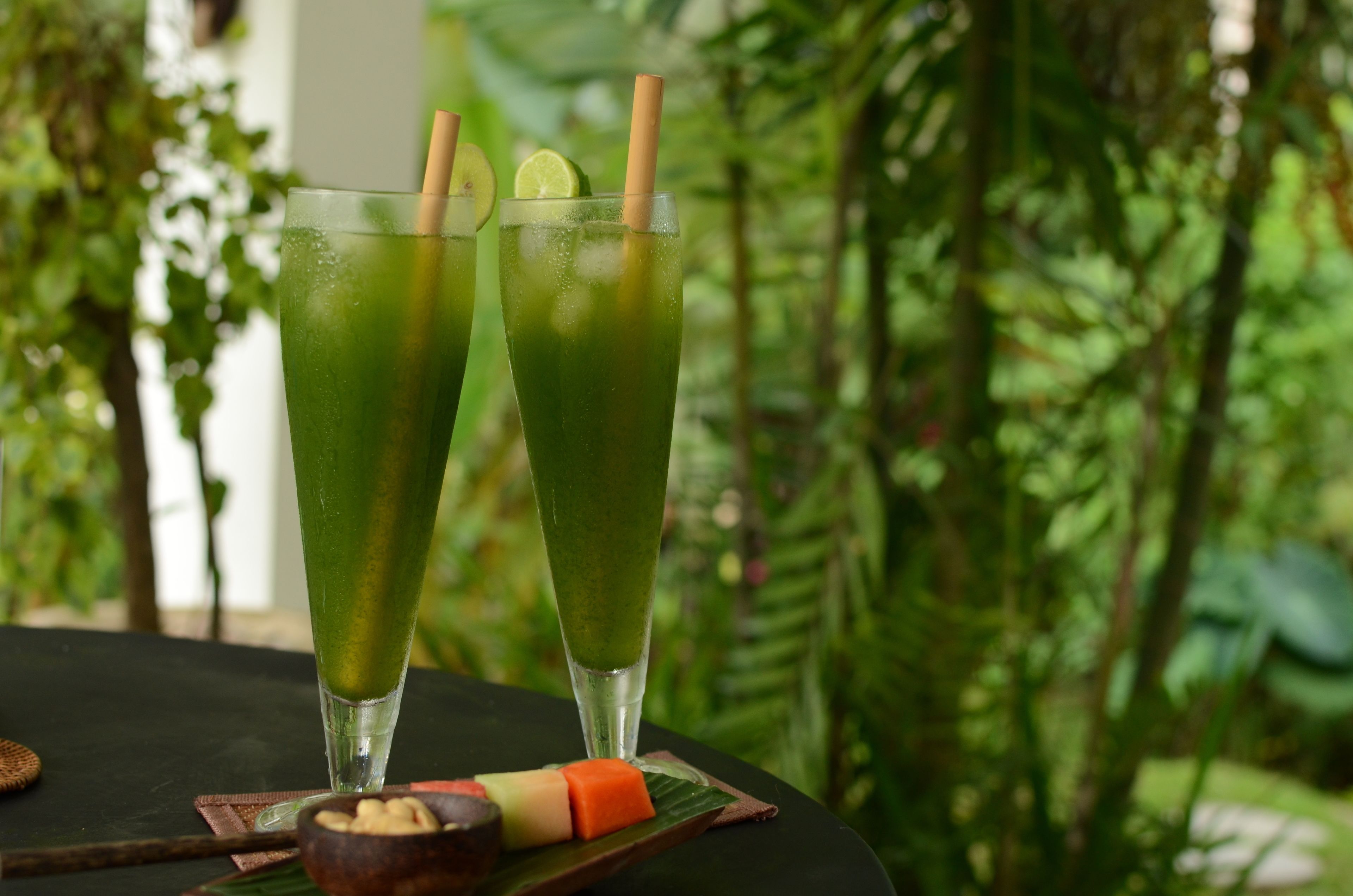 Two tall glasses of green beverage with straws and lime slices, placed on a table with a natural backdrop.