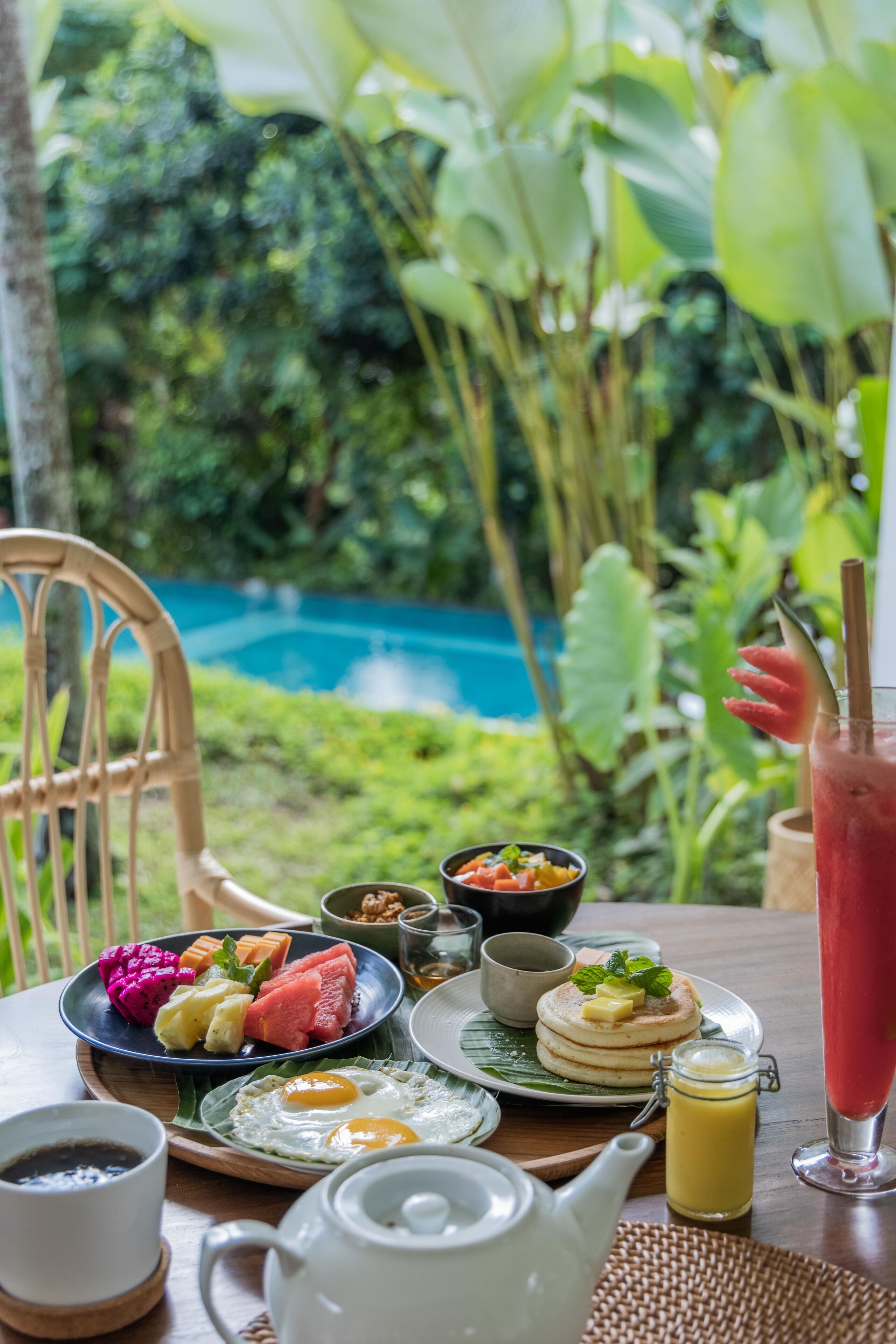 A breakfast table with various dishes, a teapot, and a drink, set against a lush garden backdrop.