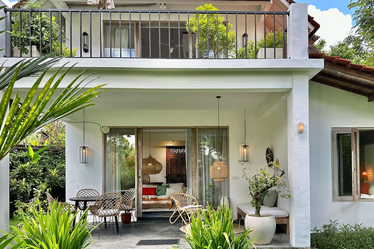 A two-story house with a balcony, a dining area with wicker furniture, and a cozy interior with a sofa and cushions.