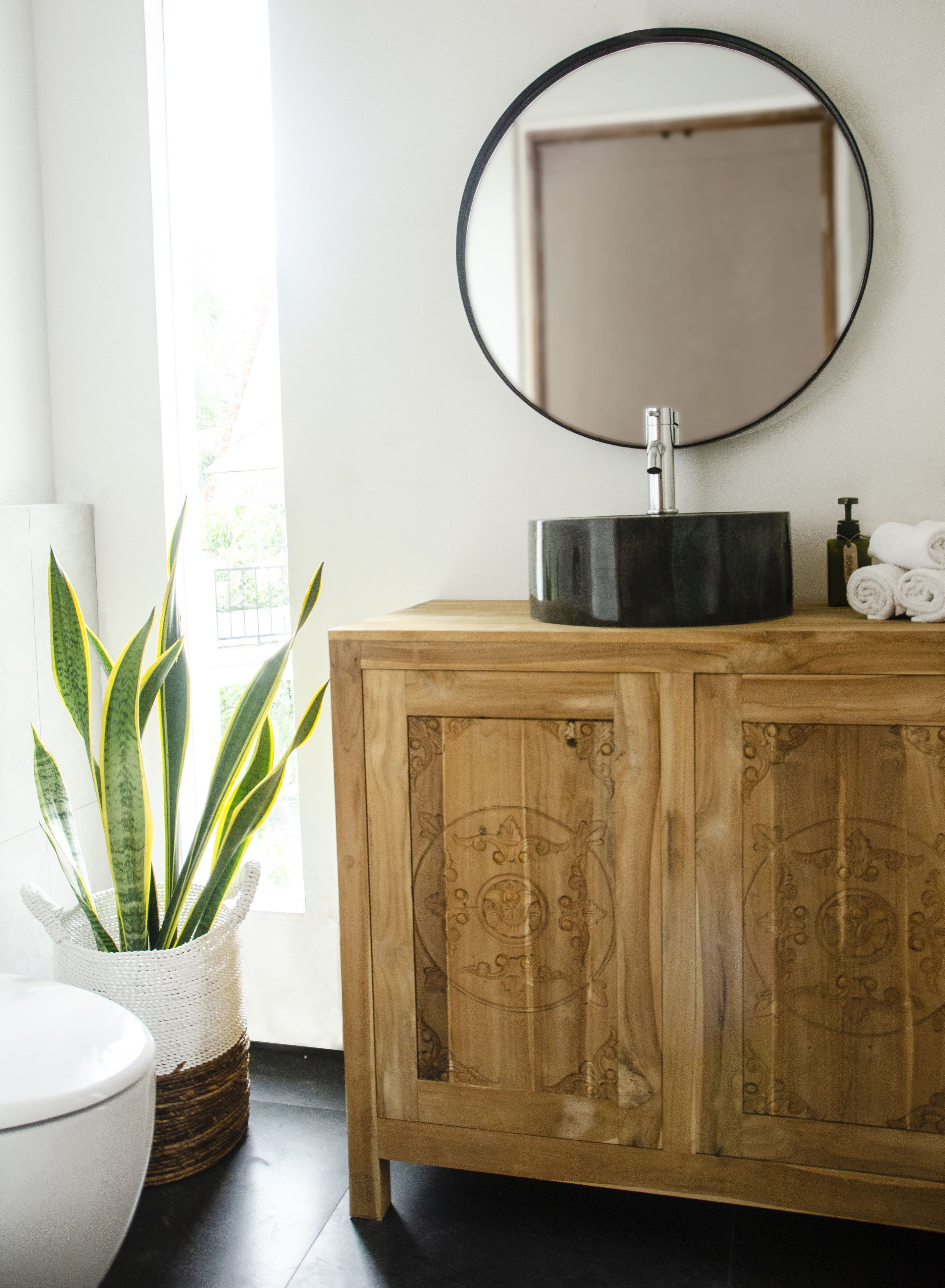 A bathroom with a wooden vanity, a round mirror, a sink, a potted plant, and a toilet.
