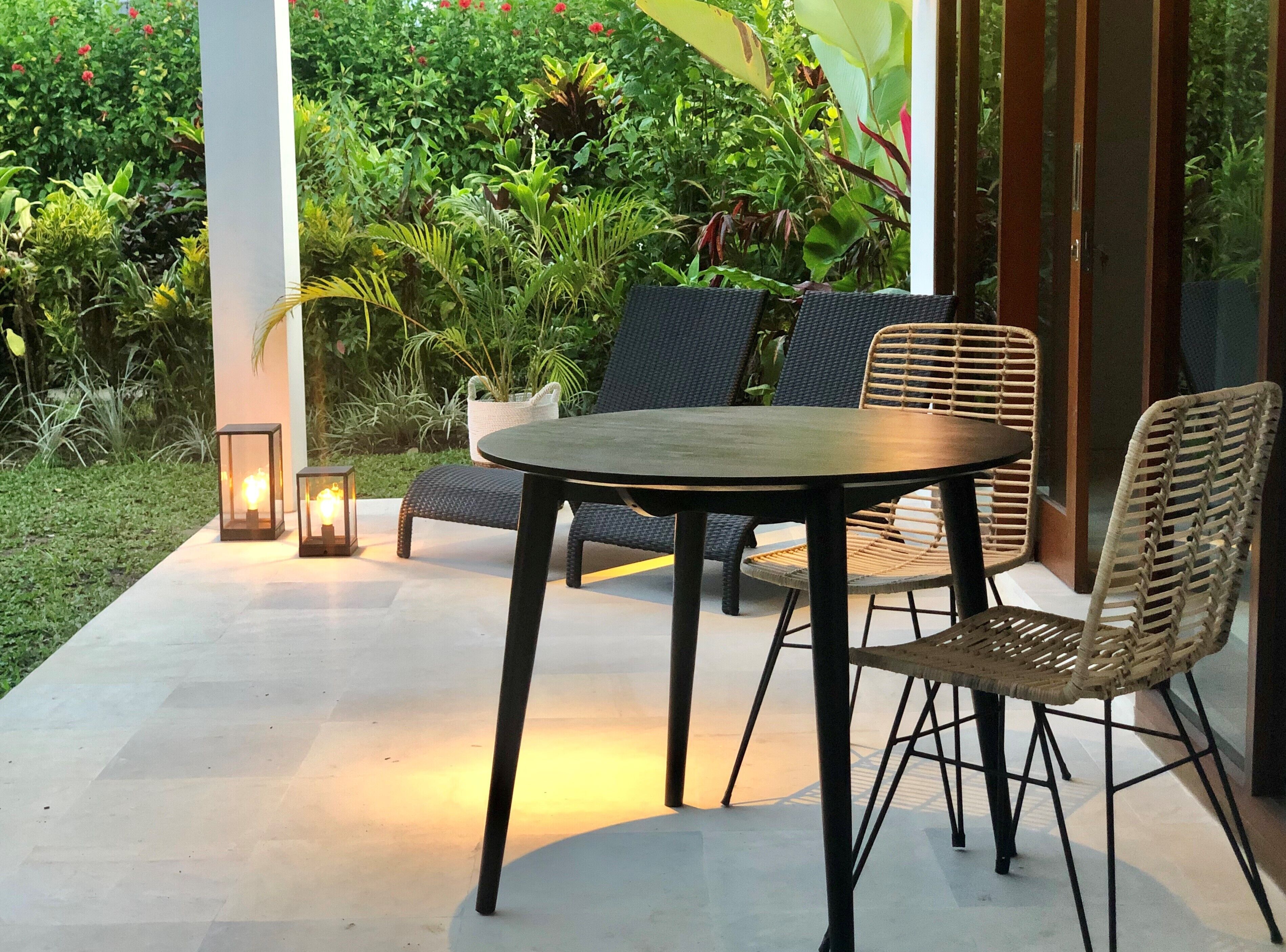 Un patio al aire libre con una mesa redonda y sillas de mimbre, rodeado de exuberante follaje y un jardín.
