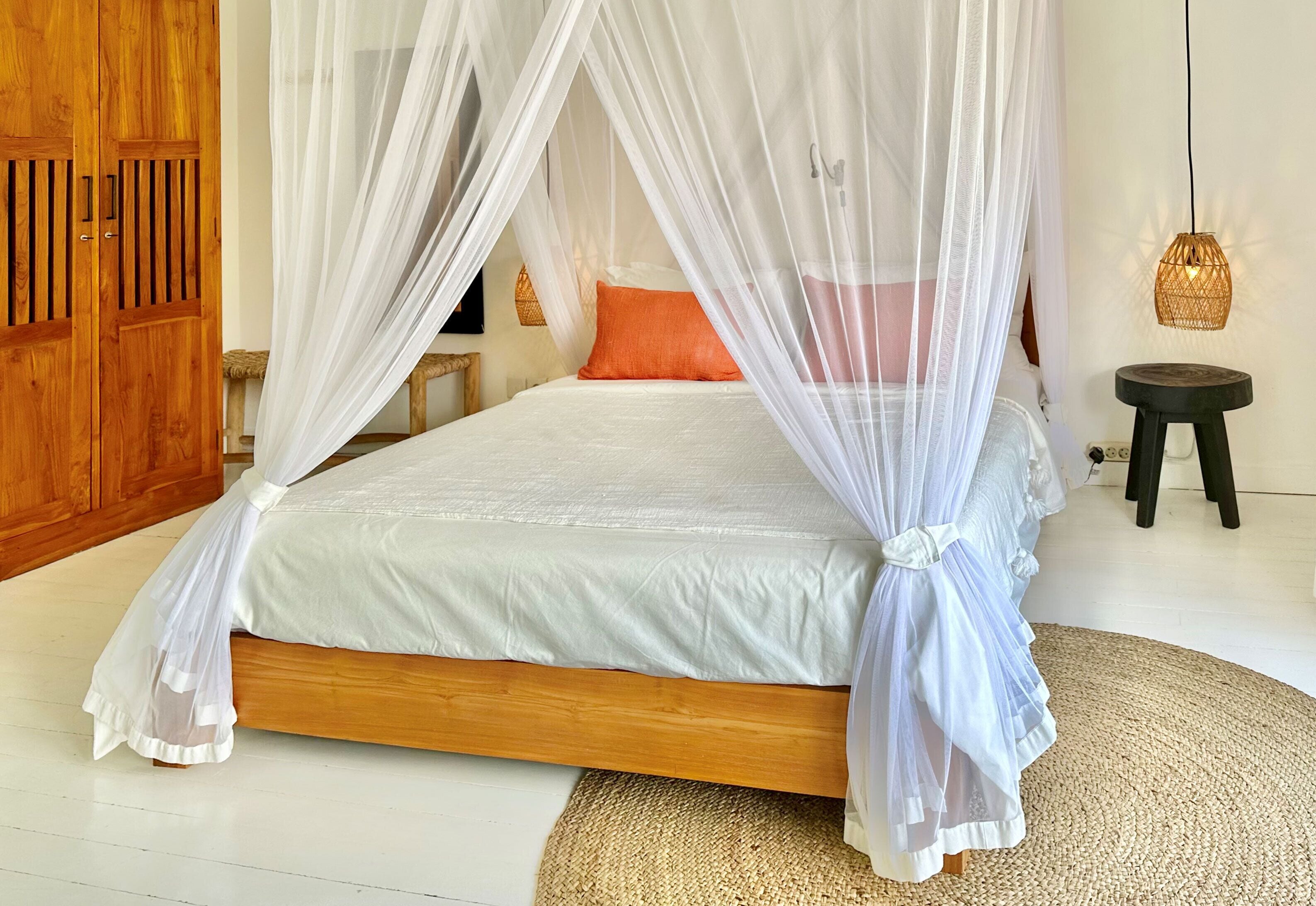 A bedroom with a bed under a white canopy, a wooden wardrobe, a wicker lamp, and a small stool.