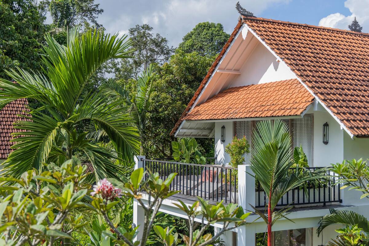 A white building with a red-tiled roof, surrounded by lush greenery and a white balcony with a black railing.
