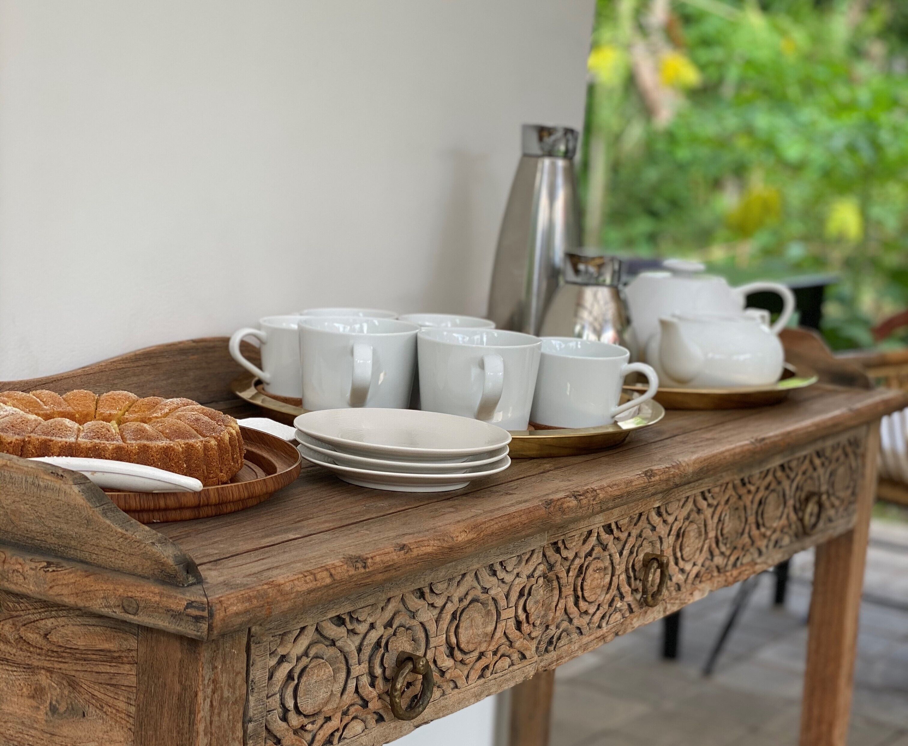 A wooden table with white cups, plates, and a teapot, set outdoors with greenery in the background.