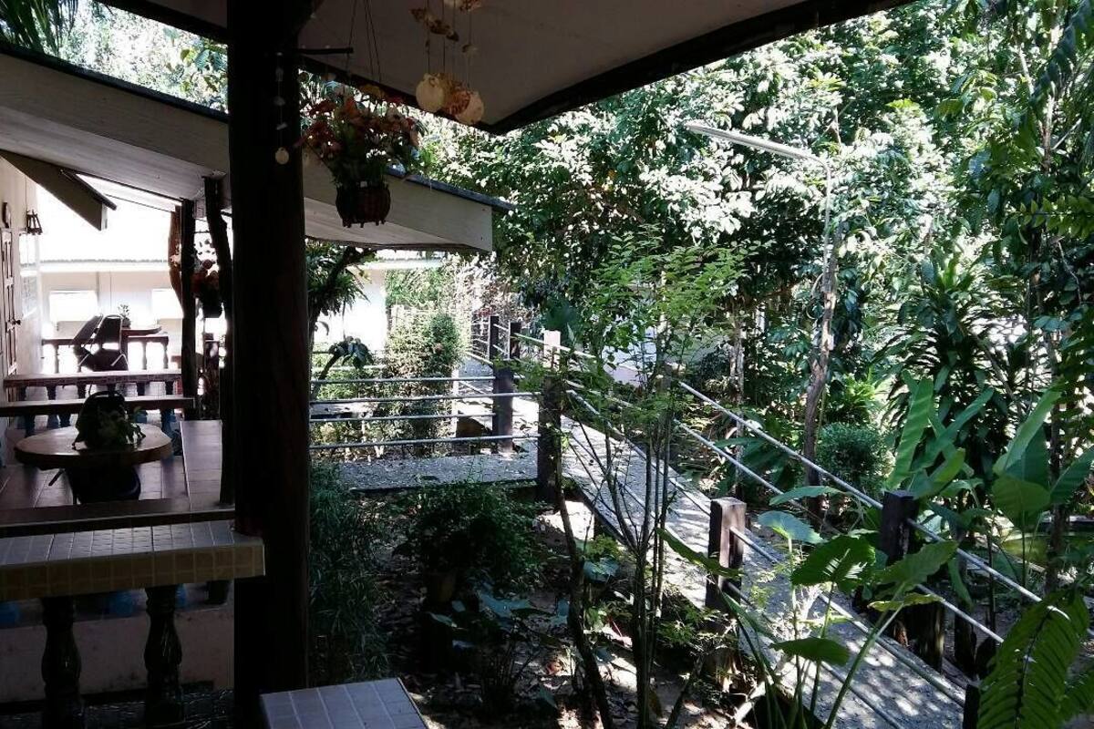 A covered outdoor area with a table and chairs, surrounded by lush greenery and a metal railing.
